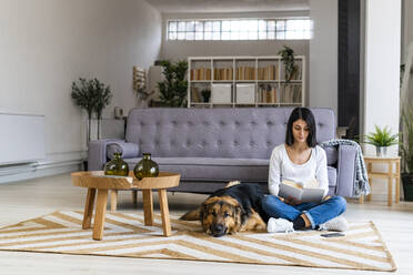 Woman reading book while sitting with dog against sofa at home - GIOF11313