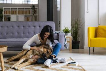 Cheerful woman embracing dog while sitting on carpet in living room - GIOF11314