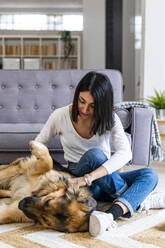 Verspielte junge Frau, die mit ihrem Hund spielt, während sie zu Hause auf dem Teppich sitzt - GIOF11315