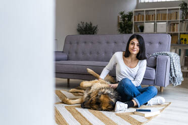 Young woman stroking pet dog while sitting on carpet against sofa in living room - GIOF11316