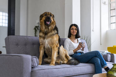 Lächelnde Frau mit Kaffeetasse, die den Hund ansieht, während sie auf dem Sofa im Wohnzimmer sitzt - GIOF11318