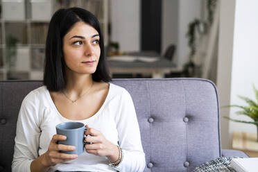 Nachdenkliche junge Frau mit Kaffeetasse im Wohnzimmer, die wegschaut - GIOF11321