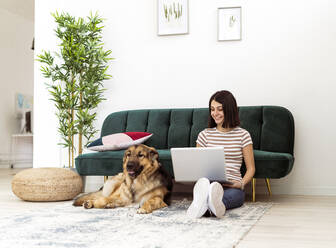 Lächelnde junge Frau mit Laptop, die einen Hund ansieht, während sie auf dem Sofa im Wohnzimmer sitzt - GIOF11324