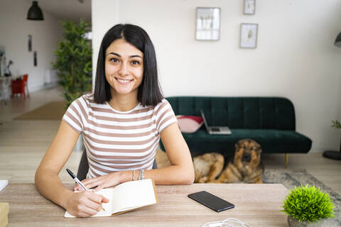 Lächelnde Frau, die einen Stift hält, während sie am Tisch im Wohnzimmer sitzt, lizenzfreies Stockfoto