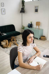 Contemplated young woman with pen and notebook sitting at table in living room - GIOF11334