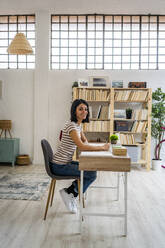 Woman sitting on chair at table against bookshelf in living room - GIOF11335