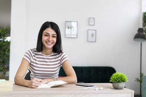 Cheerful woman with notebook and pen sitting at table in living room stock photo