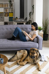 Junge Frau liest ein Buch, während der Hund zu Hause auf dem Teppich sitzt - GIOF11340
