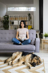 Lächelnde Frau mit Buch auf dem Sofa sitzend, während der Hund auf dem Teppich im Wohnzimmer schläft - GIOF11341