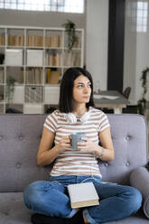 Thoughtful young woman with coffee cup sitting on sofa while looking away - GIOF11343