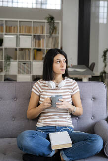 Nachdenkliche junge Frau mit Kaffeetasse auf dem Sofa sitzend und wegschauend - GIOF11343