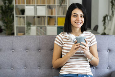 Happy woman with coffee cup and wireless headphones sitting on sofa at home - GIOF11344