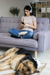 Junge Frau hört Musik, während der Hund auf dem Teppich im Wohnzimmer schläft - GIOF11345