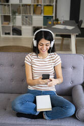 Happy woman using smart phone while listening to music through wireless headphones in living room - GIOF11347