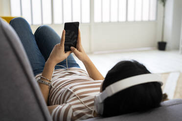 Entspannte junge Frau hört Musik über drahtlose Kopfhörer, während sie zu Hause auf dem Sofa liegt - GIOF11350