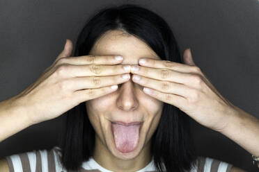 Junge Frau, die ihre Augen mit den Händen bedeckt und die Zunge herausstreckt, vor grauem Hintergrund - GIOF11359