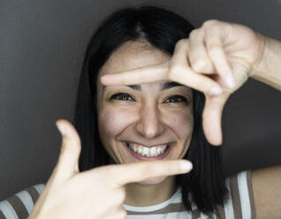 Fröhliche junge Frau schaut durch den Finger vor grauem Hintergrund - GIOF11360