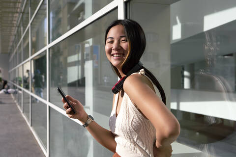 Smiling woman with smart phone and headphones around neck against glass wall stock photo