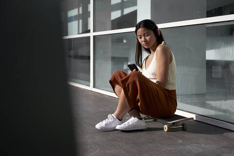Asiatische Frau, die ein Smartphone benutzt, während sie auf einem Skateboard sitzt, an einem sonnigen Tag, lizenzfreies Stockfoto