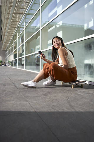 Young woman with smart phone sitting over skateboard on footpath stock photo