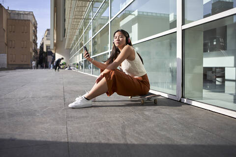 Woman with mobile phone sitting on skateboard during sunny day stock photo