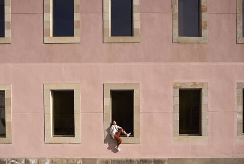 Frau sitzt auf der Fensterbank eines Gebäudes an einem sonnigen Tag, lizenzfreies Stockfoto