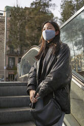 Asiatische Frau mit Handtasche, die während der Pandemie auf einer Rolltreppe steht und wegschaut - VEGF03970