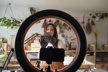 Mature female influencer preparing ring light and smart phone for video tutorial at workshop - VEGF03974