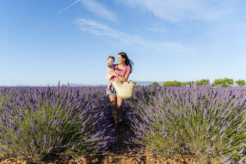 Mother carrying baby daughter in vast lavender field during summer stock photo