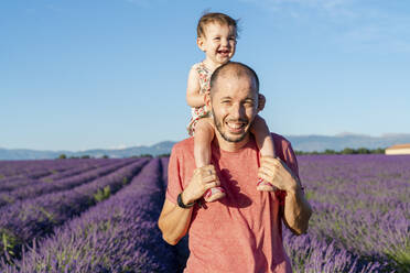 Porträt eines Vaters, der seine kleine Tochter in einem weiten Lavendelfeld huckepack nimmt - GEMF04705