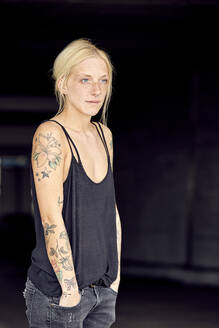 Blond woman with hands in pockets looking away while standing at parking garage - UKOF00141