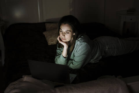 Junge Frau schaut auf einen Laptop, während sie zu Hause auf dem Bett liegt, lizenzfreies Stockfoto