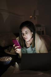 Woman with laptop and popcorn using mobile phone while lying on bed at home - AFVF08206