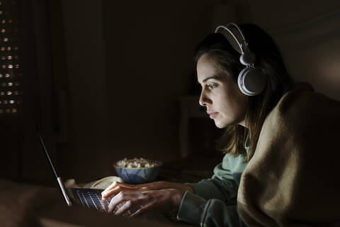 Frau mit Kopfhörern und Popcorn am Laptop, während sie zu Hause liegt, lizenzfreies Stockfoto