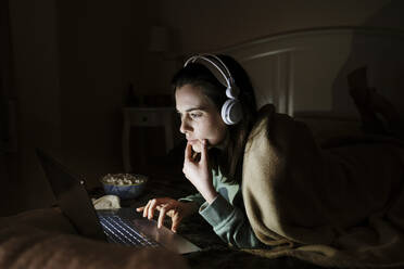 Junge Frau mit Kopfhörern schaut einen Film auf dem Laptop, während sie sich zu Hause auf dem Bett ausruht - AFVF08208