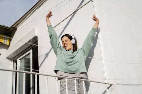 Smiling woman wearing headphones stretching hands while standing in balcony - AFVF08217