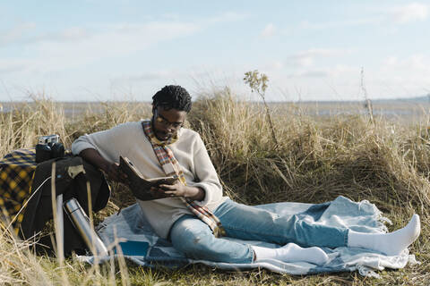 Junger Mann liest ein Buch, während er auf einer Decke am Strand sitzt, gegen den bewölkten Himmel, lizenzfreies Stockfoto