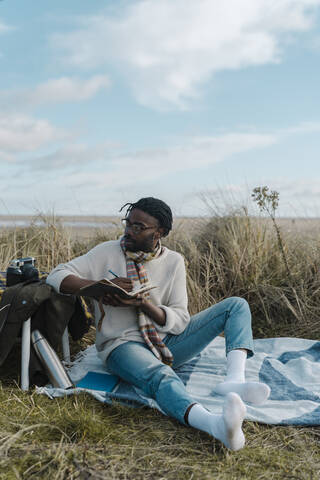 Young man with diary sitting on blanket at beach stock photo