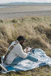 Junger Mann schreibt in Tagebuch, während er auf einer Decke inmitten von getrockneten Pflanzen am Strand sitzt - BOYF01840