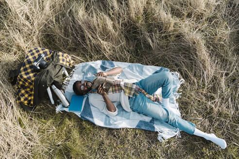 Relaxed man reading book while lying on blanket amidst dried plants - BOYF01842