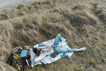 Mann mit Händen hinter dem Kopf, der sich auf einer Decke inmitten von getrockneten Pflanzen am Strand entspannt - BOYF01843