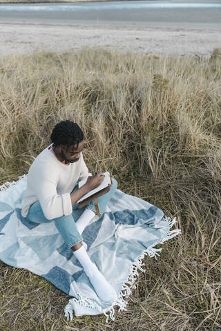 Man sitting on blanket amidst dried plant while writing in diary at beach stock photo