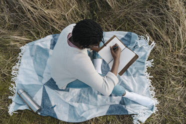 Junger Mann, der auf einer Decke sitzend inmitten von getrockneten Pflanzen Tagebuch schreibt - BOYF01848