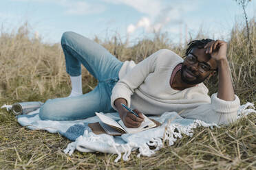 Lächelnder junger Mann mit Tagebuch auf einer Decke am Strand liegend gegen bewölkten Himmel - BOYF01849