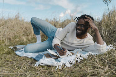 Fröhlicher junger Mann mit Tagebuch auf einer Decke am Strand liegend gegen bewölkten Himmel - BOYF01850