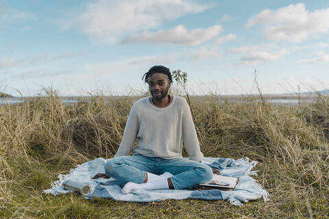 Young man sitting on blanket amidst dried plant against sky stock photo