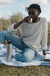 Relaxed man drinking tea while sitting on blanket against cloudy sky - BOYF01856