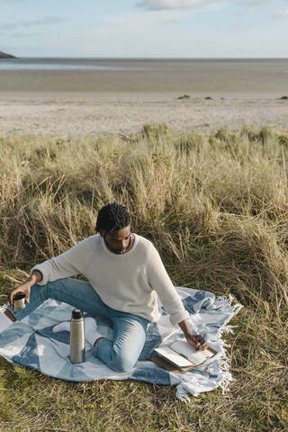 Young man holding cup while writing in diary at beach stock photo