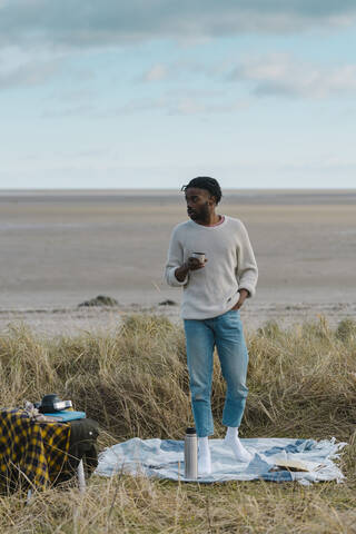 Junger Mann trinkt Tee, während er am Strand vor einem bewölkten Himmel steht, lizenzfreies Stockfoto