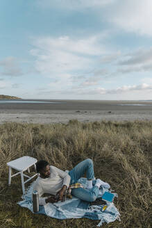 Mann benutzt sein Smartphone, während er auf einer Decke am Strand liegt - BOYF01869